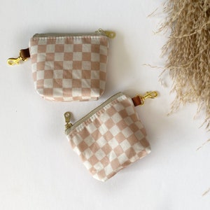 Puede incluir: Dos pequeñas bolsas de cremallera a cuadros rosa y blanco con tiradores de cremallera dorados y correas de cuero marrón.