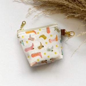 Puede incluir: Pequeño monedero blanco con cremallera y un estampado de botas de vaquero coloridas y estrellas amarillas. Tiene una cremallera dorada, una lengüeta de cuero marrón y un cierre dorado. La palabra "Howdy" está impresa.