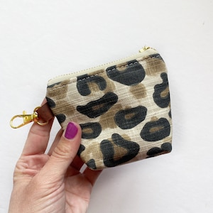 Puede incluir: Una pequeña bolsa con cremallera con un diseño de estampado de leopardo. La bolsa tiene una cremallera dorada y una correa de cuero marrón con un llavero dorado.