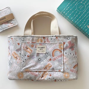 May include: A white tote bag with a cream colored fabric handle. The bag has a pocket on the front with the text "SEPTEMBER SKYE" and a floral pattern with the words "GOD'S PROMISE" and "BLESS" on the front.