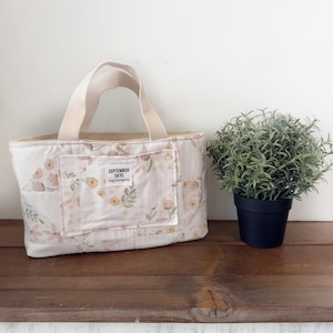 May include: A floral print tote bag with cream-colored handles and a front pocket. The bag features a pattern of pink and yellow flowers and green leaves on a white background. The text "SEPTEMBER SKYE" is on the front pocket.