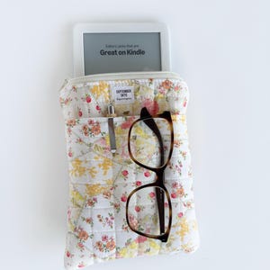 May include: A white quilted floral print case with a Kindle e-reader, glasses, and a pen. The e-reader displays "Editors' picks that are Great on Kindle." The case has a zipper closure and a label that reads "SEPTEMBER SKYE."