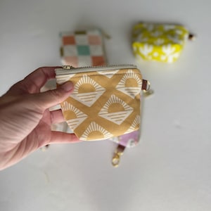 Puede incluir: Una pequeña bolsa amarilla con cremallera con un patrón geométrico blanco de soles y olas. La bolsa está en una mano.