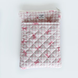 Könnte beinhalten: Eine gesteppte Tasche mit rosa-weißem Vichy-Muster und rosa Schleifenmotiven. Sie hat einen Reißverschluss und eine Vordertasche. Der Text "SEPTEMBER SKYE" ist auf einem kleinen Etikett aufgedruckt.