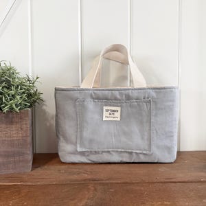 Könnte beinhalten: Hellgraue Tragetasche mit rechteckiger Tasche und cremefarbenen Griffen. Auf der Tasche steht "SEPTEMBER SKYE" auf einem kleinen Etikett. Sie steht auf einer Holzoberfläche neben einem Holztopf mit Grünpflanzen.