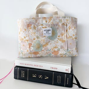 May include: A floral print tote bag with a cream-colored handle and a front pocket. The bag is resting on top of two books. The bag has a label that says "SEPTEMBER SKYE things & accessories". The books are titled "NEW MORNING MERCIES" and "ESV STUDY BIBLE".