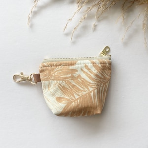 Puede incluir: Una pequeña bolsa de cremallera beige con una correa de cuero marrón y un llavero dorado. La bolsa tiene un estampado de hojas tropicales blancas y beige.