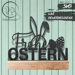 Puede incluir: Letrero de Pascua de metal negro con las palabras "Frohe OSTERN" y un diseño de orejas de conejo, montado sobre una base de madera. El letrero también incluye un diseño de huevo de Pascua. El fondo es un panel de madera verde claro.