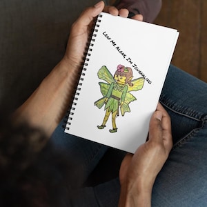 Könnte beinhalten: Weißes Spiralnotizbuch mit einer handgezeichneten Fee in Grün, mit dem Text "LEAF ME ALONE, I'M JOURNALING". Die Fee hat eine Blume im Haar und trägt einen Gürtel. Das Notizbuch wird in Händen gehalten.