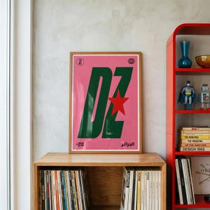 May include: A framed rectangular poster with a pink background featuring large dark green letters DZ, a red star, the number 1962, the text +213, and the word الجزائر in black. The poster is displayed on a wooden shelf above a collection of vinyl records.