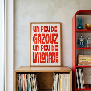May include: A framed rectangular poster with a white background and bold red typography that reads UN PEU DE GAZOUZ UN PEU DE LIMONADE. The poster is centered on a wooden shelf above a collection of vinyl records and next to a red shelving unit.