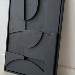 以下が含まれることがあります： グレーの色合いの、額装された抽象的な壁の芸術作品。このアートワークは、曲線や長方形を含む幾何学的な形状を持つテクスチャ加工された表面を備え、モダンな三次元効果を生み出しています。フレームは黒です。