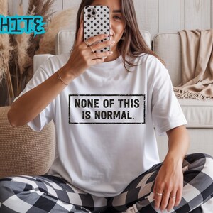 May include: White t-shirt with the text "NONE OF THIS IS NORMAL" in a black rectangular frame. The word "WHITE" is written in blue above the shirt. The person is wearing black and white checkered pajama bottoms.