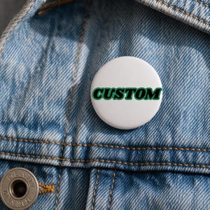 Puede incluir: Un botón blanco con la palabra "CUSTOM" en verde, contorneada en negro. El botón se encuentra sobre una chaqueta vaquera azul.