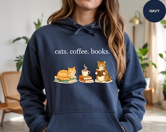 May include: Navy blue hoodie with the text "cats. coffee. books." above an illustration of two cats, a coffee cup, and books. One cat is reading a book, and the other is resting on books. The hoodie has a drawstring and a kangaroo pocket.