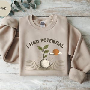 Könnte beinhalten: Ein sandfarbenes Sweatshirt mit Rundhalsausschnitt und einem Grafikdruck mit dem Text I HAD POTENTIAL. Das Design zeigt eine kleine grüne Pflanze, die aus einer runden Basis über einem aufgeschlagenen Buch wächst, sowie Sonne und Wolken.
