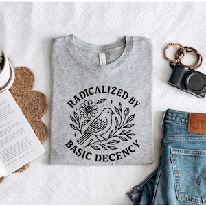 以下が含まれることがあります： 「RADICALIZED BY BASIC DECENCY」の文字と鳥と花のイラストが描かれた、グレーのTシャツ。青いジーンズ、カメラ、アクセサリーと組み合わせています。