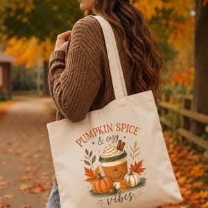 Puede incluir: Un bolso de tela color crema con un estampado gráfico de una taza de café con nata montada y ramas de canela, rodeado de hojas de otoño naranjas y verdes y calabazas, con el texto PUMPKIN SPICE & cozy vibes.