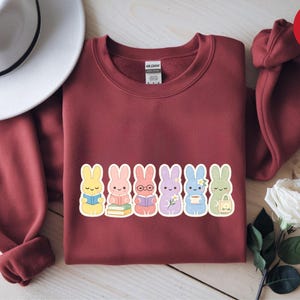 Op de afbeelding: Een bordeauxrode sweatshirt met een rij van zes cartoonkonijnen, elk met een uniek accessoire. De konijnen zijn in verschillende kleuren, waaronder geel, roze, paars, blauw en groen. De sweatshirt heeft een ronde hals en lange mouwen.