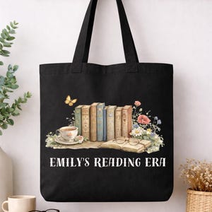 May include: Black tote bag featuring a watercolor illustration of books, flowers, a teacup, and glasses. The text "EMILY'S READING ERA" is printed in white. The bag has two black handles and is hanging on a wooden rack.