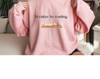 May include: A light pink crewneck sweatshirt featuring the black text 'I'd rather be reading' centered above a graphic of an open book and a tan mug resting on a wooden surface. The garment has long sleeves and a ribbed collar, cuffs, and hem.