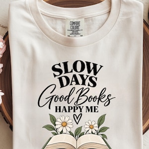 以下が含まれることがあります： アイボリー色のTシャツ。黒い文字で「SLOW DAYS Good Books HAPPY ME」と書かれ、その下に2輪の白いデイジーと緑の葉が描かれた開いた本のイラストがある。テキストと花のグラフィックの間には小さな黒いハートが配置されている。