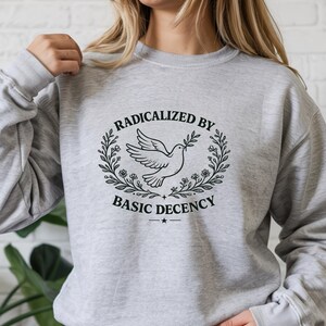 May include: A sport grey crewneck sweatshirt featuring a black graphic print of a dove carrying an olive branch, surrounded by a floral wreath. The text RADICALIZED BY BASIC DECENCY is printed in black capital letters above and below the dove.