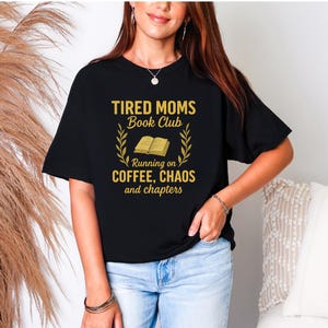 Może przedstawiać: Czarny t-shirt ze złotym napisem "TIRED MOMS Book Club" nad grafiką otwartej książki i słowami "Running on COFFEE, CHAOS and chapters". Koszulka ma swobodny styl.
