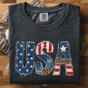 May include: A dark grey t-shirt with the word "USA" appliquéd on the front. The letters are made of denim, leopard print, and red and white stripes, with stars on the "U" and "A". The shirt is folded on a wooden surface.