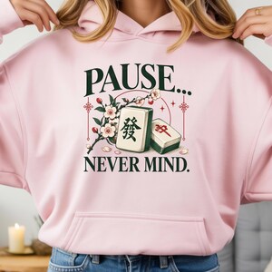 Könnte beinhalten: Ein hellrosa Kapuzenpullover mit einem Grafikdruck und dem Text PAUSE... NEVER MIND. Das Design zeigt zwei Mahjong-Steine, einen mit einem grünen und einen mit einem roten Schriftzeichen, umgeben von einem blühenden Zweig und roten Akzenten.
