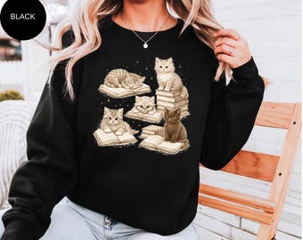 Op de afbeelding: Zwarte sweatshirt met een afbeelding van katten die boeken lezen. De illustratie toont verschillende katten, sommige zitten op stapels boeken, andere liggen. Eén kat draagt een bril. Het woord "BLACK" staat in een zwarte cirkel linksboven.