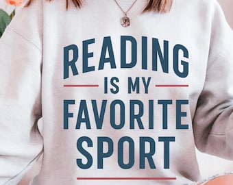 May include: A light gray sweatshirt with the text "READING IS MY FAVORITE SPORT" in navy blue block letters. The text is separated by red lines. The sweatshirt is a casual style.