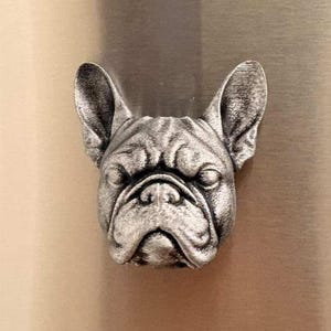 May include: A silver-colored, three-dimensional bulldog head. The sculpture has detailed features, including wrinkles and prominent ears. The bulldog's face is centered, with a slightly furrowed brow. The object is likely a decorative item or a magnet.