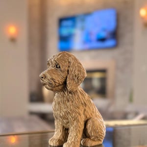 May include: A detailed, golden-brown dog figurine in a seated position. The sculpture has a textured surface, capturing the fur's appearance. The dog's ears are floppy, and its gaze is directed to the side. The background is blurred, suggesting an indoor setting.