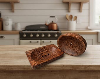 May include: Two wooden soap dishes with a natural wood grain pattern. One is rectangular with a recessed area and drainage holes, and the other is a matching oval-shaped lid. The dishes are on a wooden surface.