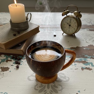 Puede incluir: Una taza humeante de café con arte latte sobre una mesa de madera envejecida, junto a una vela encendida en un soporte de metal, un reloj despertador antiguo y libros viejos. Granos de café esparcidos.