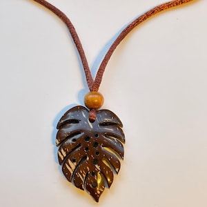 Puede incluir: Un collar con colgante en forma de hoja marrón. El colgante está hecho de un material marrón oscuro con un patrón natural y texturizado. El collar tiene un cordón marrón y una cuenta de madera. El diseño recuerda al follaje tropical.
