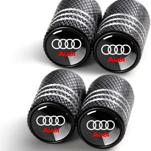 May include: Set of four black tire valve stem caps with a textured design. Each cap features a white Audi logo and the word "Audi" in red. The caps have a silver band and are designed for car tires.