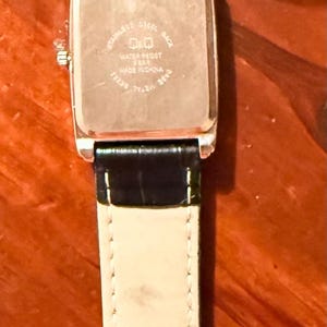 K&ouml;nnte beinhalten: Eine Q&Q-Armbanduhr mit einem rechteckigen, silberfarbenen Zifferblatt und einem schwarzen Lederarmband. Die Uhr hat einen Edelstahlboden und einen Schnallenverschluss. Das Armband ist 20 cm lang.