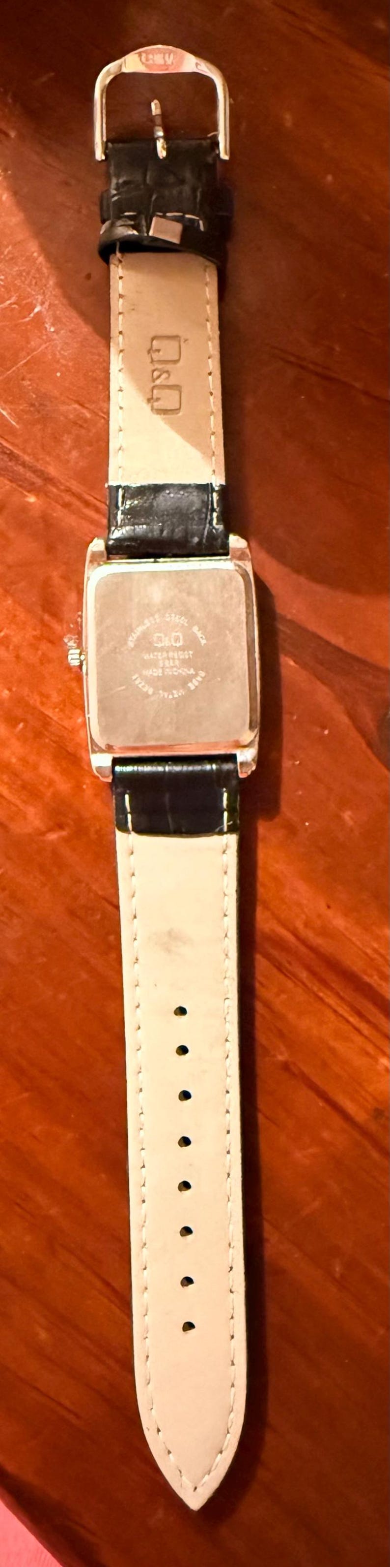 K&ouml;nnte beinhalten: Eine Q&Q-Armbanduhr mit einem rechteckigen, silberfarbenen Zifferblatt und einem schwarzen Lederarmband. Die Uhr hat einen Edelstahlboden und einen Schnallenverschluss. Das Armband ist 20 cm lang.