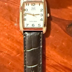 K&ouml;nnte beinhalten: Eine rechteckige Q&Q Quartz-Uhr mit schwarzem Lederarmband. Das Zifferblatt ist wei&szlig; mit schwarzen Ziffern und Zeigern. Das Armband ist etwa 20 cm lang.