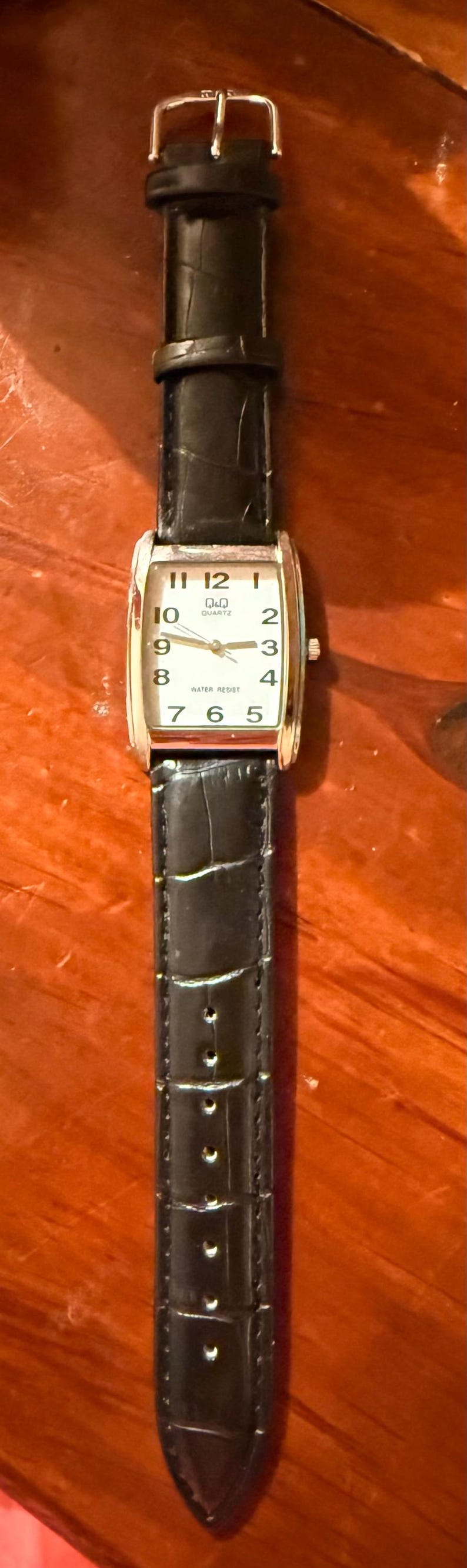 K&ouml;nnte beinhalten: Eine rechteckige Q&Q Quartz-Uhr mit schwarzem Lederarmband. Das Zifferblatt ist wei&szlig; mit schwarzen Ziffern und Zeigern. Das Armband ist etwa 20 cm lang.