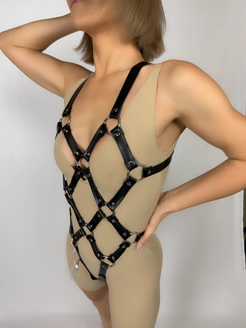May include: Black leather body harness with a geometric design. The harness features a series of interconnected straps and metal rings. The design covers the chest and torso, with straps extending to the hips.