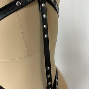 May include: Black leather harness with silver-tone metal rivets. The harness features multiple straps and rings, designed to be worn over clothing. The design includes a series of vertical straps with evenly spaced rivets.