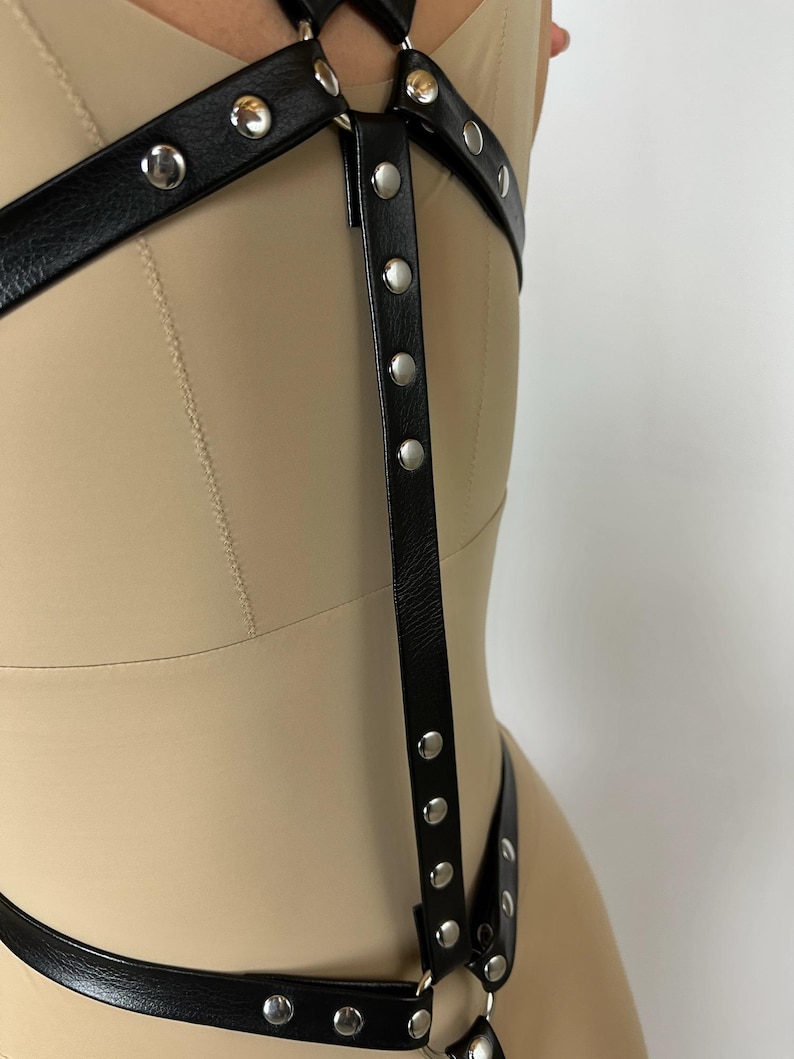 May include: Black leather harness with silver-tone metal rivets. The harness features multiple straps and rings, designed to be worn over clothing. The design includes a series of vertical straps with evenly spaced rivets.