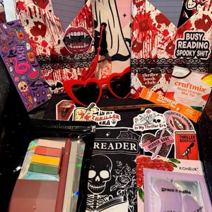 May include: A collection of spooky themed items, including heart-shaped sunglasses, stickers, and a book sleeve with a skeleton design. The items are arranged in a black container, with the text "BITE ME" and "BUSY READING SPOOKY SHIT".