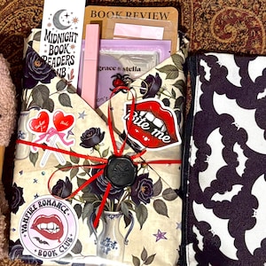 May include: A gift set featuring fuzzy beige socks with a heart design, a book wrapped in floral paper with a black wax seal, and a black and white bat-patterned pouch. The package includes stickers reading "Bite me" and "Vampire Romance Book Club."