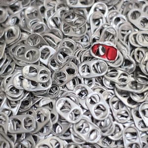 以下が含まれることがあります： 銀色のアルミ缶のプルタブの山をクローズアップ。1つのタブは赤く塗られています。タブは散らばっており、質感のある金属の表面を作り出しています。タブには2つの穴と湾曲したハンドルが付いています。