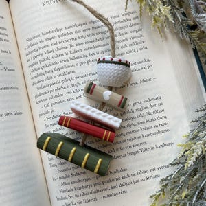 Puede incluir: Una colecci&oacute;n de accesorios para libros hechos a mano, que incluyen varios marcadores en miniatura con forma de libro en rojo, verde y blanco, y un peque&ntilde;o cuenco blanco de ganchillo con ribete verde y rojo, todo sobre un libro abierto.