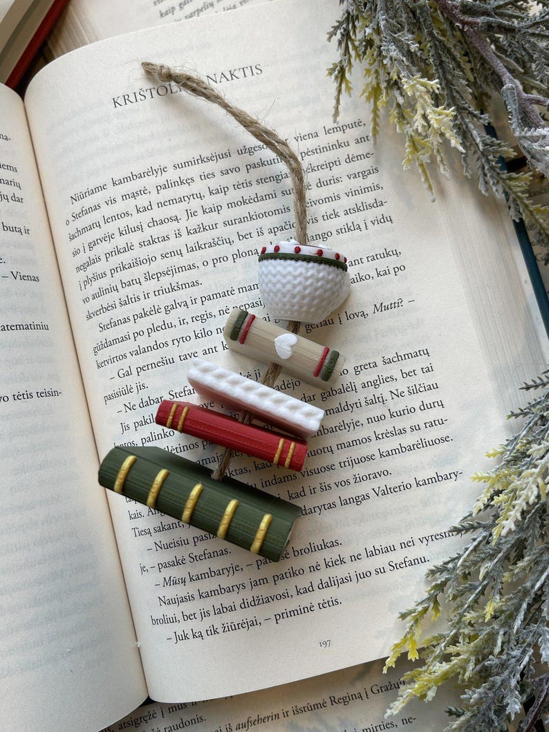 Puede incluir: Una colecci&oacute;n de accesorios para libros hechos a mano, que incluyen varios marcadores en miniatura con forma de libro en rojo, verde y blanco, y un peque&ntilde;o cuenco blanco de ganchillo con ribete verde y rojo, todo sobre un libro abierto.